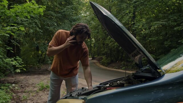 Driver stands beside the open hood of a broken-down car on a rural roadside and talks on the phone about the malfunction. He may be consulting an insurance company or an auto mechanic.