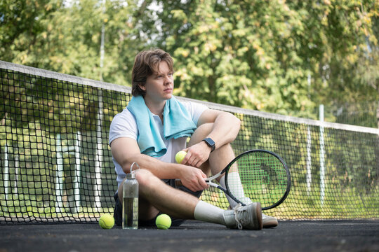 Young male tennis player sitting beside net with racket, balls and water bottle after workout, representing rest, recovery and sports fatigue.