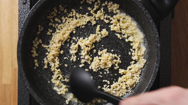 Silicone spoon stirs chopped almonds in frying pan. Low heat toasts almonds evenly without burnt taste or dark spots. Students stir almonds, learning heat control for golden colour and crisp texture.