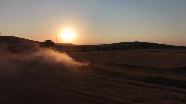 Golden hour landscape of a harvested farm field with agricultural machinery creating dust.