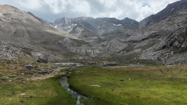 Cinematic forward tracking shot over a green mountain stream towards rugged rocky peaks under a cloudy sky.