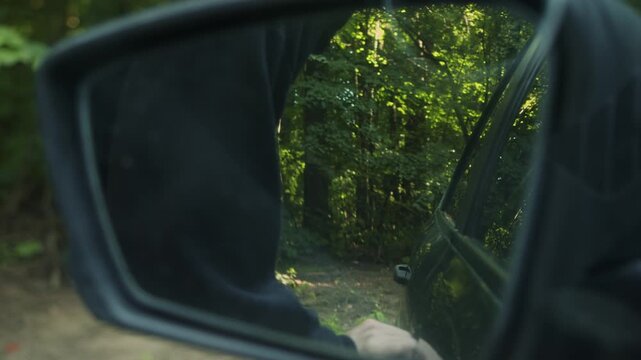View through side mirror shows masked thief breaking car door lock on forest roadside. Criminal forces entry into vehicle and steals car.