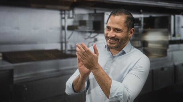 Man clapping hands and smiling while standing by stainless steel ovens in a commercial kitchen building; team spirit joy.