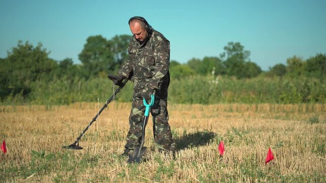 Male treasure hunter in camouflage outfit uses metal detector while holding a shovel in a field marked with red flags, surrounded by greenery under a clear blue sky
