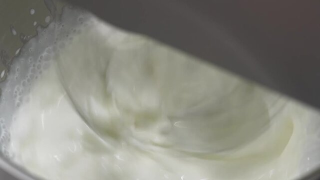 close-up shot of a metal wire whisk attachment on a stand mixer whipping heavy cream or a milky mixture in a bowl, preparing a base for a delicious fruit dessert.
