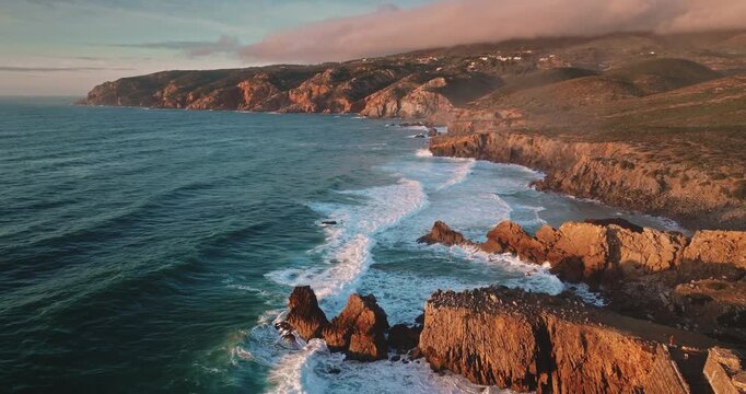 Sintra Cascais Natural Park aerial panorama, rugged coastline with powerful ocean waves breaking against rocky cliffs and shore under golden hour sunset light. Travel and explore Portugal nature