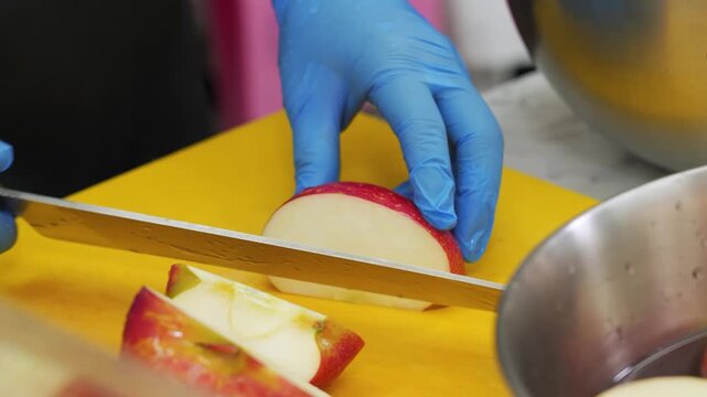 Close-up of a chef wearing blue gloves using a sharp knife to slice fresh red apples on a bright yellow cutting board. The professional food preparation focuses on creating a fresh fruit dessert in a