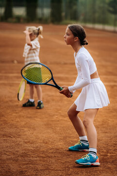 Two children practice tennis on an outdoor clay court, holding rackets in their hands and concentrating on a sports training exercise.