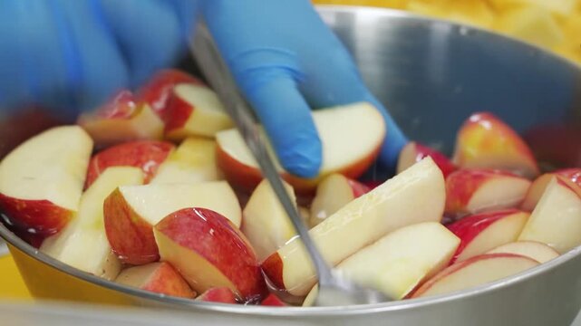 Close-up footage of crisp red apple slices soaking in water inside a stainless steel bowl, prepared for a fruit dessert or baking recipe in a bright kitchen setting.