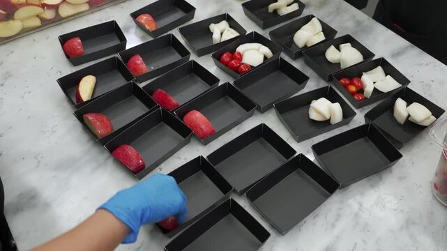 High-angle view of a chef wearing blue gloves arranging sliced apples, pears, and strawberries into neat rows of black square dessert trays on a marble countertop in a professional catering kitchen.