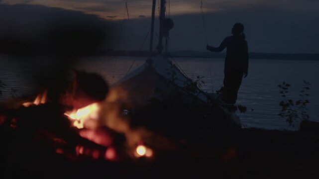 Sailing and camping in the wild. Woman sailor walks on the wild area with sailing boat on the background and bonfire on the foreground