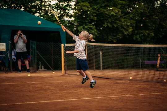 Little boy hitting tennis ball over net in jump on outdoor clay court, active childhood and dynamic sports training.