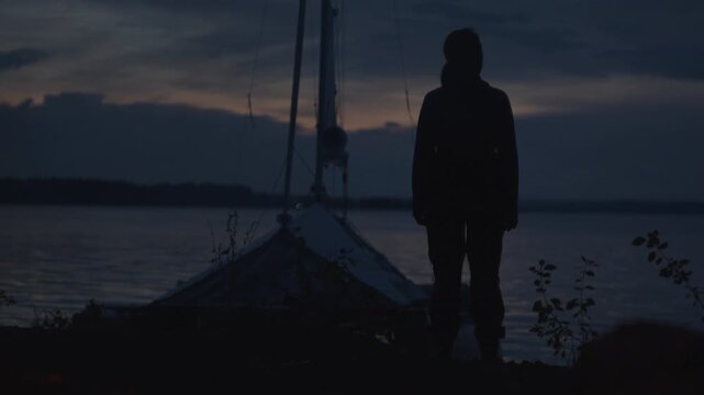 Sailing and camping in the wild. Woman sailor walks on the wild area with sailing boat on the background and bonfire on the foreground