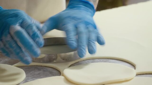 Close-up of a pastry chef wearing blue protective gloves using a metal ring mold cutter to stamp out perfect circles from rolled out white dough on a professional kitchen counter.