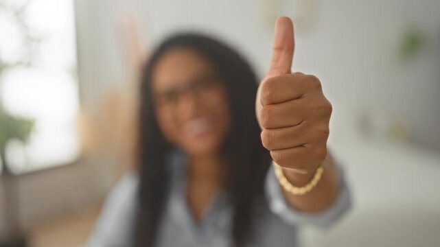 Woman wearing glasses smiling and showing thumb up hand with chunky gold bracelet in a studio setting; confidence success empowerment.