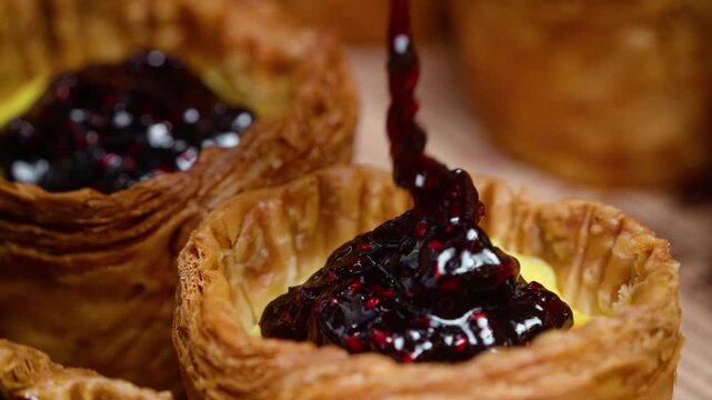 Close up video of dark sweet berry jam or compote being poured onto a golden, flaky puff pastry cup filled with creamy yellow vanilla custard. Perfect for bakery advertisements, dessert recipes, and f