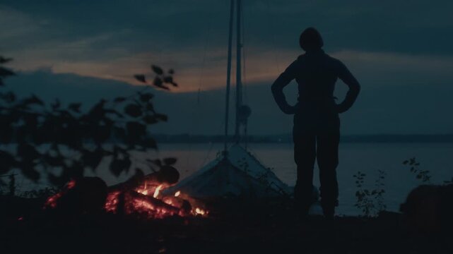 Sailing and camping in the wild. Woman sailor walks on the wild area with sailing boat on the background and bonfire on the foreground