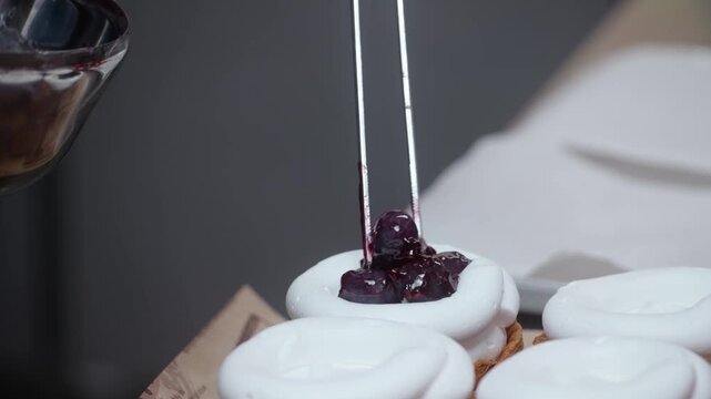 Close up of a confectioner using a spoon to add rich, glossy cherry fruit sauce topping onto white, whipped meringue tartlets or pavlovas on a commercial kitchen workbench.