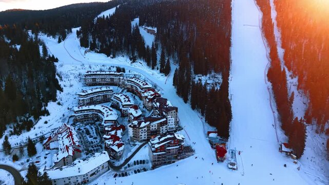 Aerial view of a mountain resort village surrounded by snow. Buildings with dark roofs sit near steep ski tracks illuminated by warm sunlight.