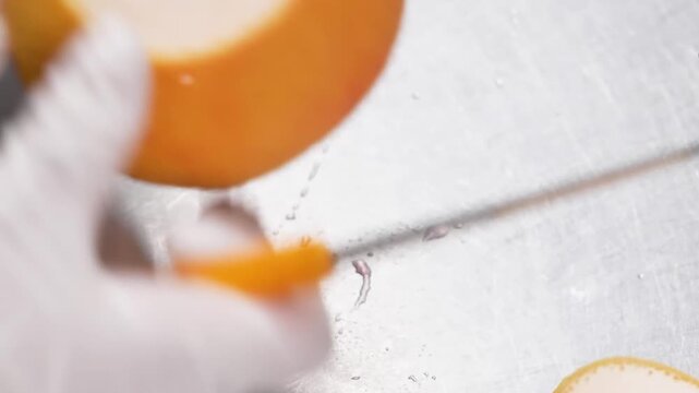 Close-up video of a pastry chef wearing white gloves meticulously cutting and preparing a fresh, juicy ruby red grapefruit on a professional stainless steel kitchen counter to make a gourmet fruit des