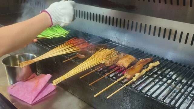 A chef grills skewers of marinated meat, mushrooms, and other ingredients over charcoal on a commercial barbecue grill, with smoke rising from the fire
