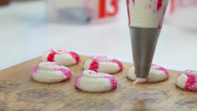 Close-up of a pastry chef using a piping bag with a metal nozzle to squeeze out white and red striped macaron batter onto a wooden pattern baking surface