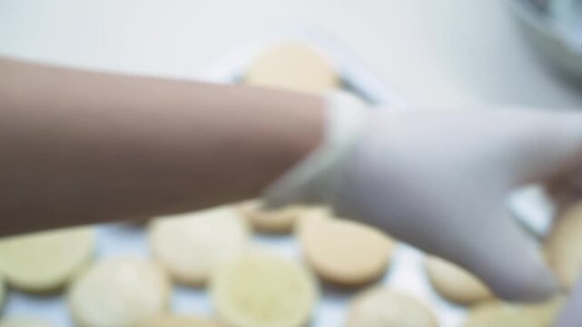 A pastry chef wearing protective white nitrile gloves uses a pastry brush to apply a delicate glaze or coating to round macaron shells in a commercial kitchen setup.