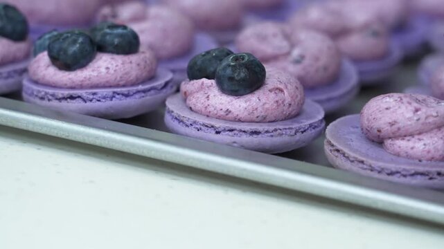 Close-up shot of purple macaron shells arranged on a metal baking tray, piped with smooth berry buttercream and topped with fresh blueberries during the baking process