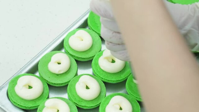 Close-up of a pastry chef wearing gloves using a piping bag to squeeze white cream or ganache onto bright green macaron shells. The sweet French meringue-based confections are laid out on a baking tra