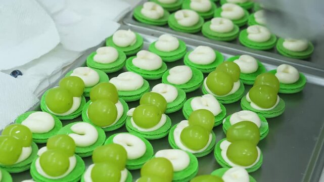 Close-up high-angle view of rows of bright green macaron shells being assembled on a metal baking sheet. Each dessert is topped with pipeable white cream filling and fresh, juicy green grapes, showcas