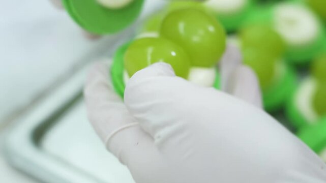 Close-up shot of a pastry chef wearing white hygiene gloves carefully assembling a bright green macaron dessert. The delicate French pastry is filled with smooth white cream filling and stuffed with f