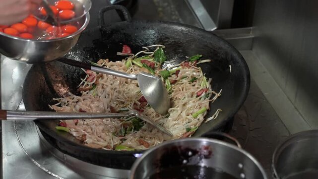Chef stir-frying flat rice noodles with vegetables and bean sprouts in a hot wok, showing authentic Asian cooking in a restaurant kitchen.