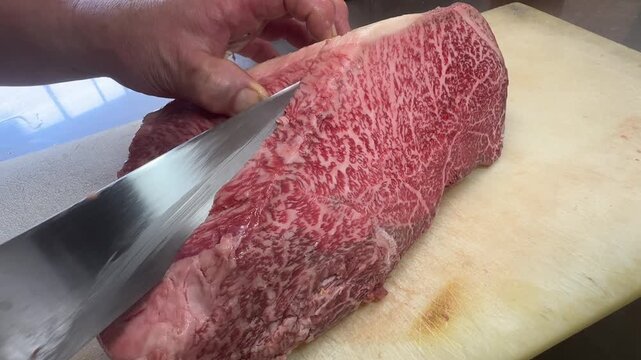 Chef slicing a raw premium Japanese wagyu beef block into thick marbled steaks with a knife on a white cutting board, close-up, Japan