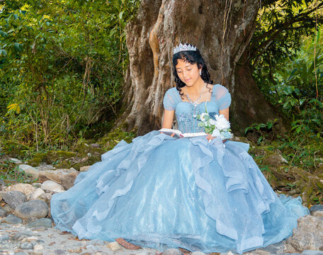 bajo un &aacute;rbol y lee un libro. Feliz Quincea&ntilde;era con Corona Leyendo un Libro en un Entorno Natural. Quincea&ntilde;era Sonriente Sosteniendo un Libro en un Hermoso Escenario Natural.
