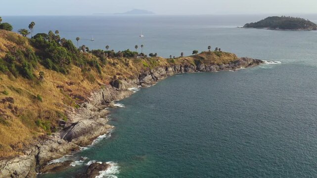 Promthep Cape coastal landscape with a lush green and dry grass hill, rocky cliffs meeting turquoise ocean waves, and distant tropical islands under clear sky, serene view of Phuket island. Drone shot