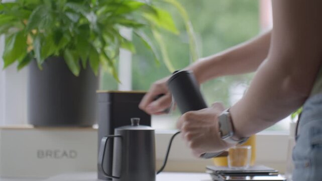 Woman grinds coffee with manual hand grinder beside plant and glass cup on kitchen counter. Concept of coffee preparation and fresh brewing