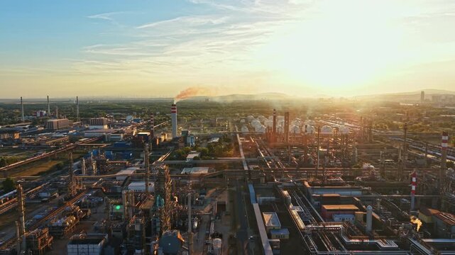 Workers operate machinery at an oil refinery located outside the city during sunset. Smoke rises from the stacks as the sun sets in the background.