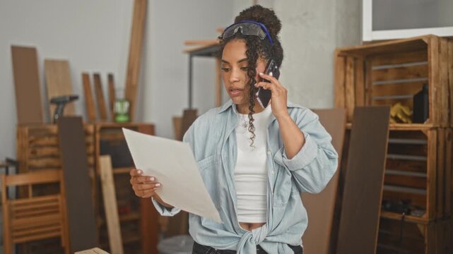 Woman carpenter holding smartphone to ear and reading a sheet of plans in studio workshop amid wooden planks and crates; concentration planning.
