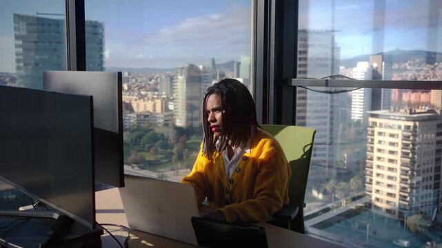 Focused Black businesswoman typing on a laptop while colleagues work and talk in the background. Sunlit corporate scene conveying concentration, productivity and responsibility