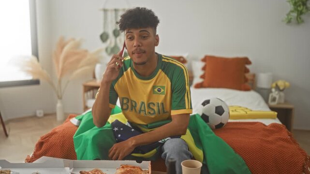 Man in brazil jersey holding smartphone to ear with right hand and reaching for pizza on bed in bedroom; excitement support.