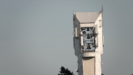 Nowoczesna, biała wieża kościelna z zamontowanymi dzwonami, na której umieszczono także współczesne anteny i nadajniki telekomunikacyjne na tle czystego nieba. © tomo81