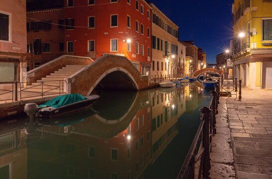 Quiet canal with moored boats, brick bridges and colorful buildings reflected in calm water before sunrise in Venice, Italy.