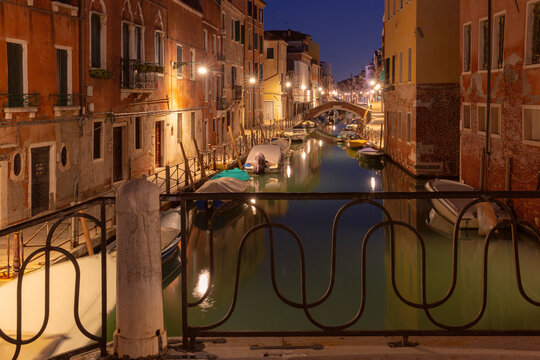 Quiet canal with moored boats, brick bridges and colorful buildings reflected in calm water before sunrise in Venice, Italy.
