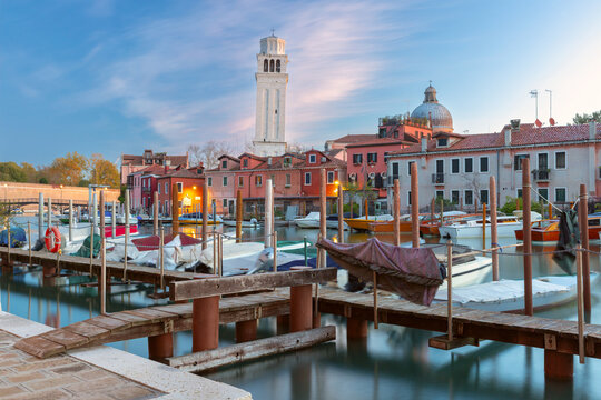 Boats moored along wooden piers beside traditional buildings and a bell tower at dawn in the Castello district of Venice, Italy.