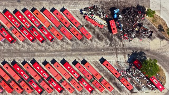 Long rows of red buses are parked in a scrapyard. Nearby, there are piles of metal and wrecked vehicles. The scene shows a busy place where transportation vehicles are stored.