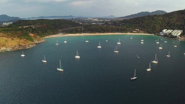 Amazing Nai Harn Bay aerial panorama. Numerous sailboats and yachts moored in the calm, turquoise waters of Phuket island coastline with sandy beach and lush green headland, Thailand. Drone shot