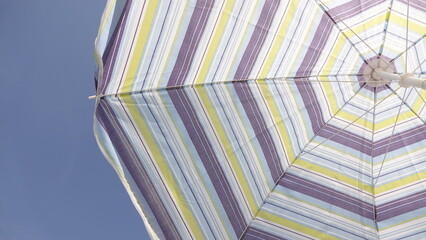 Widok od dołu na rozłożony parasol ogrodowy w kolorowe paski. Zdjęcie ukazuje geometryczną, symetryczną konstrukcję stelaża zbiegającą się w centralnym punkcie. © tomo81