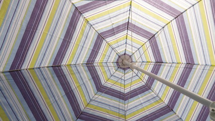 Widok od dołu na rozłożony parasol ogrodowy w kolorowe paski. Zdjęcie ukazuje geometryczną, symetryczną konstrukcję stelaża zbiegającą się w centralnym punkcie. © tomo81