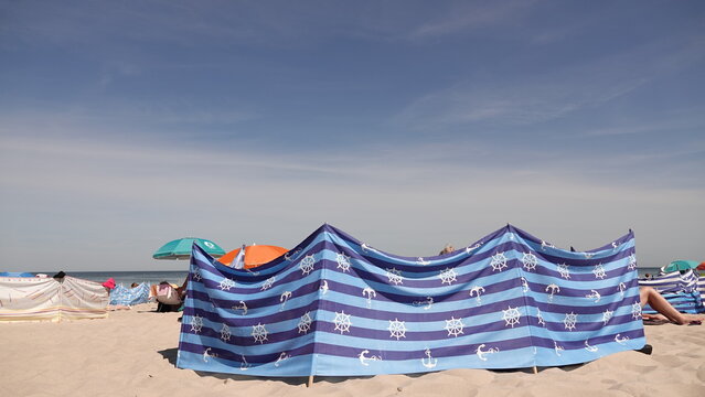 Zdjęcie przedstawia rozłożony na piaszczystej plaży niebiesko-granatowy parawan w paski z motywem kotwic i ster&oacute;w. W tle widać czyste, bezchmurne niebo.