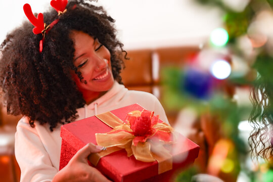 Two happy multiethnic young women hold a red present tied with a gold bow in a cosy home decorated for Christmas surrounded by holiday decorations and warm light